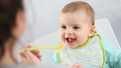 Frau füttert Baby mit Brei. Das Baby trägt ein Lätzchen und freut sich.