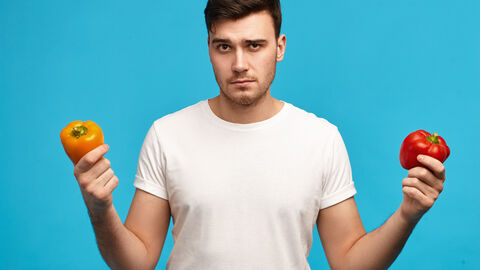 Mann hält zwei Paprika in jeweils einer Hand und schaut kritisch