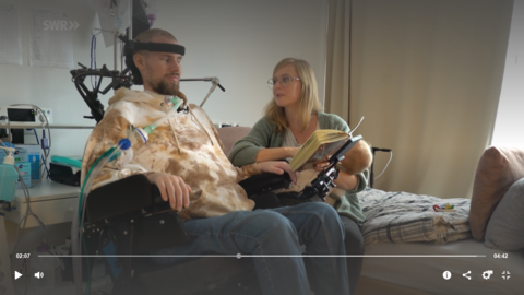 Es wird eine Szene aus dem Beitrag des SWR "Gelähmt und beatmet: Jan infiziert sich mit dem Rattenbiss-Fieber" gezeigt. Die Schwester hält ein Buch so, dass Jan daraus lesen kann-