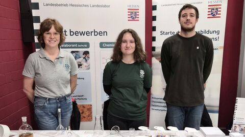 Drei Auszubildene stehen vor Rolls-Up-Displays und hinter einem Tisch.