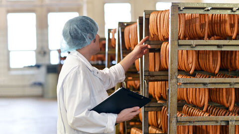 Ein Lebensmittelkontrolleur befindet sich in einem Lagwerbereich eines Wurstherstellers und weist mit einer Hand auf eine Reihe von Würstchen, die auf einem Gestell aufgehängt sind.