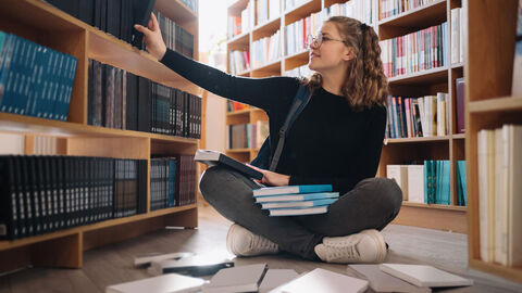 Eine junge Frau sitzt in einer Bibliothek vor einem Regal mit Büchern und zieht ein einzelnes buch aus einem Regalfach. 