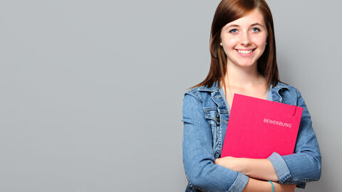 Eine junge Frau steht vor einem grauen Hintergrund und hält einen Berwerbungsmappe vor ihren verschrenkten Armen.